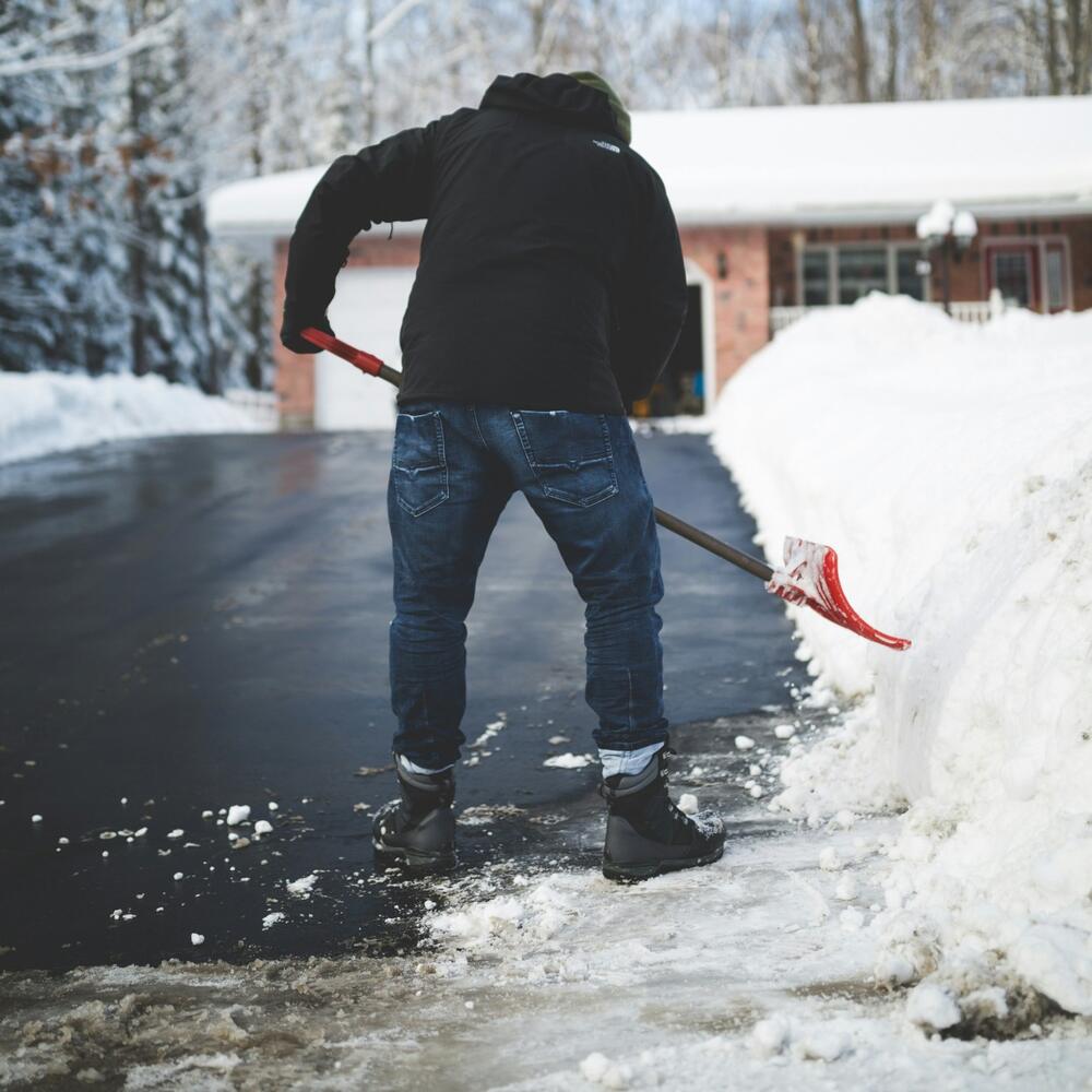 Person shoveling a cleared asphalt driveway next to a tall bank of snow